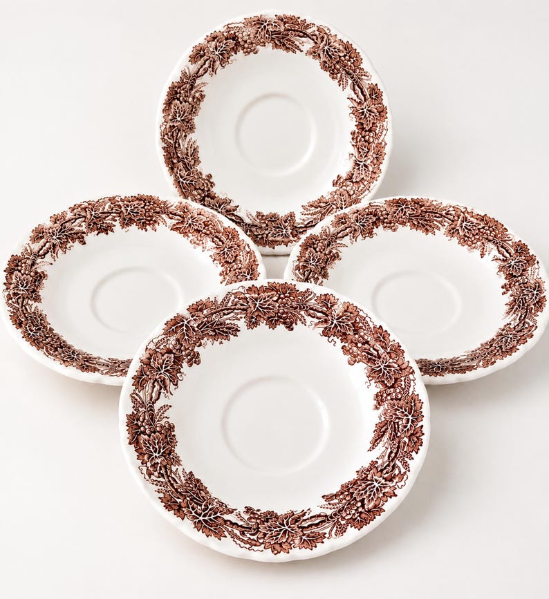 May include: A set of four white ceramic saucers, each featuring a brown transferware pattern along the rim. The design consists of detailed illustrations of grape clusters and vine leaves arranged in a continuous circular border around the center.