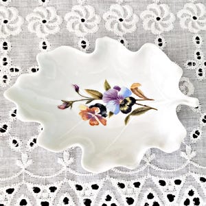 May include: White leaf-shaped dish with a floral design. The dish features a painted arrangement of purple, blue, and orange pansies with green leaves. The dish is approximately 8 inches long and 6 inches wide.