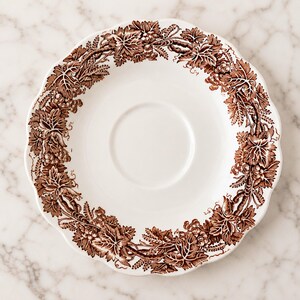 May include: A white ceramic saucer featuring a scalloped edge and a brown transferware pattern along the rim. The decorative border displays a detailed botanical motif of intertwined grapevines, clusters of grapes, and wheat stalks in brown.