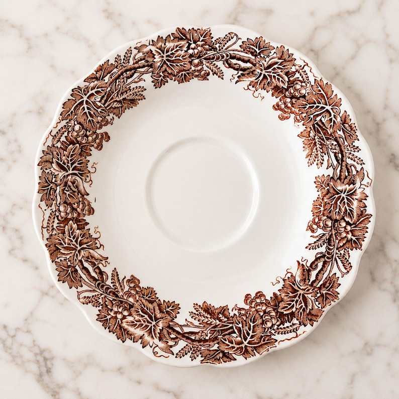 May include: A white ceramic saucer featuring a scalloped edge and a brown transferware pattern along the rim. The decorative border displays a detailed botanical motif of intertwined grapevines, clusters of grapes, and wheat stalks in brown.