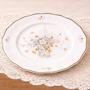 Puede incluir: Plato de cerámica blanca con borde festoneado y borde dorado. El plato presenta un diseño floral en colores pastel, incluyendo rosa, azul y amarillo. El plato está sobre un mantel de encaje.