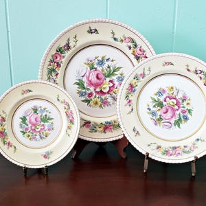 May include: Three antique decorative plates with floral designs. The plates are cream-colored with a gold rim and a beaded edge. The center of each plate features a bouquet of pink, blue, and yellow flowers. The plates are displayed on stands.
