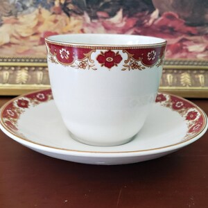 May include: A white porcelain teacup and matching saucer set featuring a decorative border pattern in deep red and gold. The rim of the cup and the edge of the saucer are adorned with a repeating floral motif in red with gold accents and thin lines.