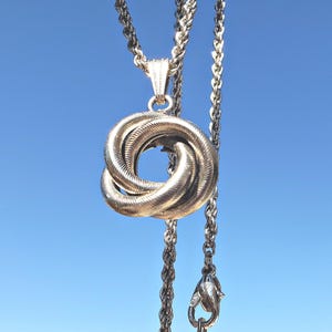 May include: Silver necklace featuring a pendant in the form of a knotted design. The chain is a rope style, and the pendant has a textured finish. The backdrop is a clear blue sky.