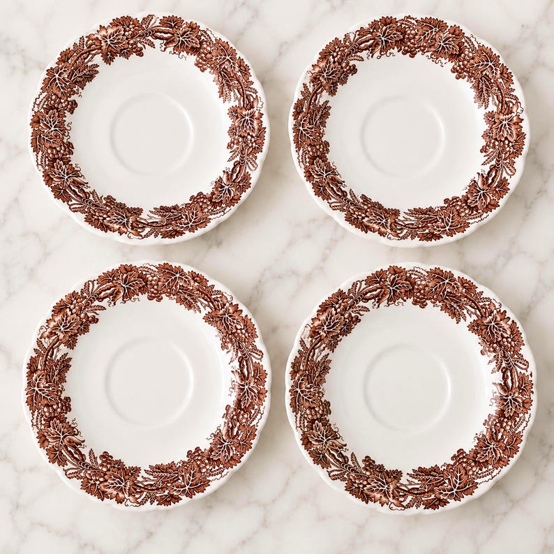 May include: A set of four round white ceramic saucers, each featuring a detailed brown transferware pattern along the rim. The design consists of intricate grapevines, leaves, and clusters of grapes encircling the center of each saucer.