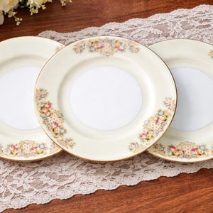 May include: Three cream-coloured ceramic plates with floral designs and gold rims. The plates have a white centre and are arranged on a lace tablecloth. The floral pattern includes pink, yellow, and blue flowers.