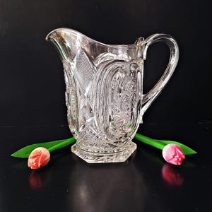 May include: A clear, vintage-style glass pitcher with intricate embossed patterns. The pitcher features a spout, a handle, and a hexagonal base. Two tulips with pink and red petals and green stems are placed on either side of the pitcher.