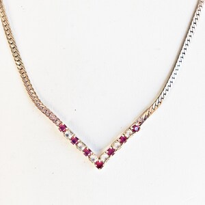 Può includere: Una collana color oro con un pendente a forma di V. Il pendente è adornato con pietre preziose rosse e trasparenti alternate. La catena ha un design piatto e intrecciato. La collana è mostrata su sfondo bianco.