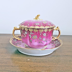 May include: A vintage pink porcelain soup bowl with a lid, handles, and saucer. The bowl and lid are decorated with floral patterns and gold trim. The set is on a wooden surface.