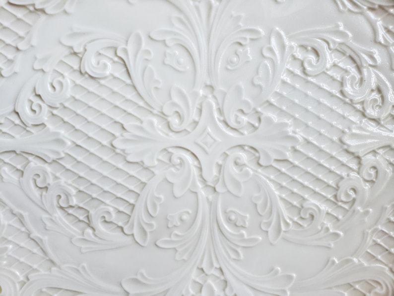 May include: White ceramic plate with a raised, embossed design of intricate floral patterns. The design is symmetrical and features a repeating pattern of leaves and swirls.