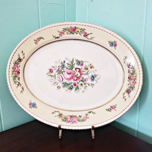 May include: Oval serving platter with a cream-colored background and a floral design. The platter features a central bouquet of pink roses and blue flowers, with additional floral accents around the rim. A gold trim borders the edge.