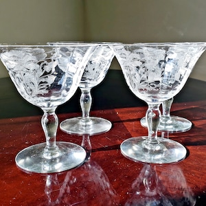 Op de afbeelding: Vier heldere glazen met gegraveerde bloemmotieven. De glazen hebben een brede, ondiepe kom en een ronde voet. Het gegraveerde bloemmotief is zichtbaar op de kom van elk glas. De glazen zijn gerangschikt op een donker houten oppervlak.