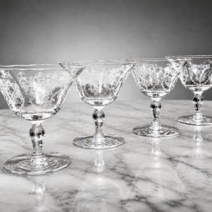 Op de afbeelding: Vier heldere cocktailglazen met geëtste bloemmotieven. De glazen hebben een brede, ondiepe kom en een gedetailleerde steel. Ze zijn gerangschikt op een marmeren oppervlak, dat het licht reflecteert. De achtergrond is een grijze gradiënt.