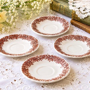 May include: A set of four white ceramic saucers, each featuring a circular indentation in the center and a decorative border of intricate brown leaf patterns around the rim. The saucers have a smooth, glossy finish and a consistent round shape.