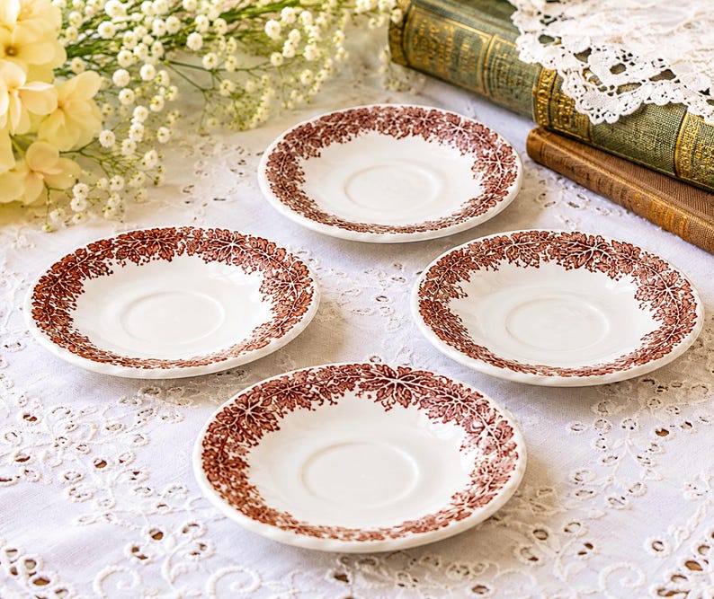May include: A set of four white ceramic saucers, each featuring a circular indentation in the center and a decorative border of intricate brown leaf patterns around the rim. The saucers have a smooth, glossy finish and a consistent round shape.