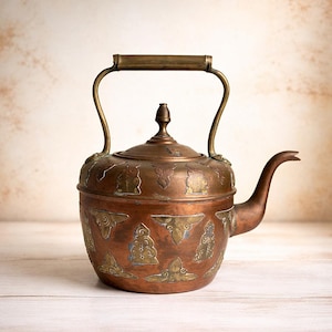 Könnte beinhalten: Antike Kupferkanne mit Messinggriff und Ausguss. Die Teekanne hat dekorative Messingakzente und einen Deckel mit einem kleinen Knauf. Der Körper der Teekanne ist mit geätzten Designs verziert.