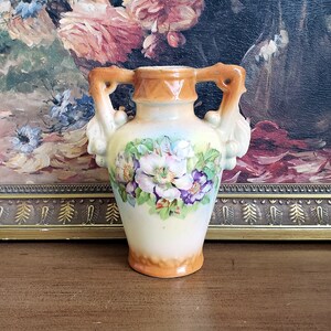 May include: A small, antique vase with a cream-colored base and orange accents. The vase features two ornate handles and is decorated with a floral design in shades of purple, white, and green. The background includes a painting with floral elements.