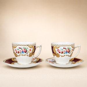 May include: Two matching teacups and saucers. The white cups have a burgundy band and gold trim. Floral designs in pink, blue, and yellow decorate the cups and saucers. The saucers have a burgundy border with gold accents.