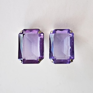 May include: Two rectangular, faceted, purple gemstone earrings with silver-colored metal accents. The earrings are set against a white background, showcasing their vibrant color and geometric shape. The gemstones have a slight gradient of color.