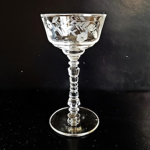 Puede incluir: Una copa de champán de cristal transparente grabado con un diseño floral. La copa presenta una copa ancha, un tallo decorativo y una base circular. El patrón floral es visible alrededor del borde del cristal.