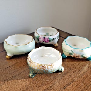 May include: Four small, decorative porcelain bowls with scalloped edges and three small feet. The bowls are painted in various colors, including green, pink, and gold, with floral and patterned designs. The bowls are arranged on a wooden surface.