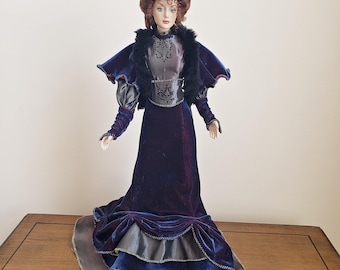 May include: A porcelain doll in a Victorian-era gown. The doll has auburn hair styled in an updo and is wearing a dark blue velvet dress with a black fur-trimmed cape. The dress features a layered skirt with gray and blue accents.