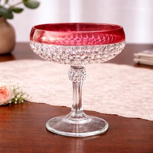 Puede incluir: Una copa de cristal transparente con borde rojo rubí. El cuenco tiene un patrón tallado en diamante y el tallo está elegantemente diseñado. La copa se asienta sobre una base redonda, perfecta para servir cócteles o postres. El vaso se exhibe sobre una superficie de madera.