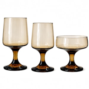 May include: Three pieces of amber-colored glassware with a smooth finish. The set includes two tall, stemmed glasses with cylindrical bowls and one wide, shallow stemmed coupe glass, all featuring a deep brown base and stem.