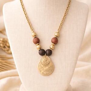 May include: Gold-toned necklace with a chain and pendant. The necklace features a series of brown and gold beads, leading to a layered, circular pendant with etched designs. The necklace is displayed on a cream-colored mannequin.