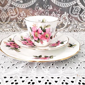 Peut inclure: Service à thé en porcelaine blanche avec une tasse, une soucoupe et une assiette. Le service est orné de motifs floraux roses et de bordures dorées. La tasse a une anse incurvée. L'ensemble est posé sur une nappe en dentelle blanche.
