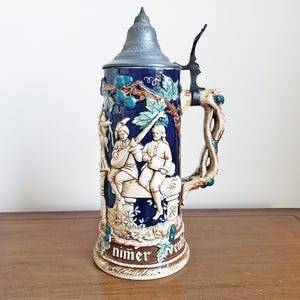 Op de afbeelding: Een keramische bierpul met een blauwe achtergrond en gedetailleerde reliëfsnijwerken. De pul heeft een tinnen deksel en handvat, met een decoratief handvat in de vorm van een wijnstok. Het reliëf toont figuren in historische kleding. De basis luidt "nimer".