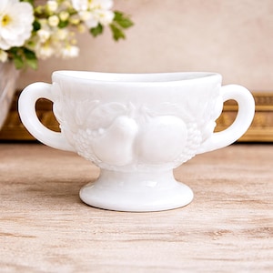 Puede incluir: Un cuenco de vidrio lechoso blanco con dos asas, con un diseño de frutas en relieve de peras, uvas y otras frutas. El cuenco se asienta sobre una base de pedestal, mostrando una estética vintage. El cuenco mide unos 10 cm de alto.