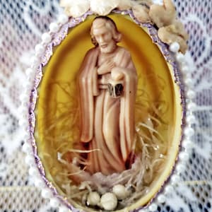May include: An egg-shaped religious ornament. Inside is a figurine of a man in robes, holding a lantern. The egg is yellow with a purple and white beaded border, and is decorated with small, white pearls and dried flowers. A decorative item.