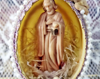Puede incluir: Adorno religioso en forma de huevo. El interior presenta una figura de un hombre con túnica, sosteniendo una linterna. El huevo es amarillo con un borde de cuentas moradas y blancas, y está decorado con pequeñas perlas blancas y flores secas.