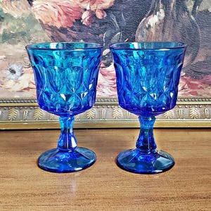 Può includere: Due calici blu vintage con un motivo a diamante. Le vetrerie sono in vetro e hanno una base rotonda. I calici sono esposti su una superficie di legno con un dipinto incorniciato sullo sfondo.