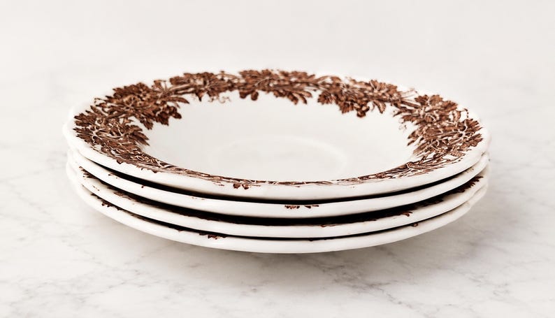 May include: A stack of three white ceramic plates featuring a brown transferware pattern of grapevines and leaves along the rim. The plates have a shallow, rounded bowl shape with a smooth, glossy finish and a detailed botanical border design.
