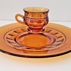 Puede incluir: Juego de taza y platillo de té de vidrio color ámbar. La taza tiene un asa y se asienta sobre un platillo a juego con un borde decorativo. El juego está hecho de vidrio transparente, mostrando su contenido. El diseño es clásico.