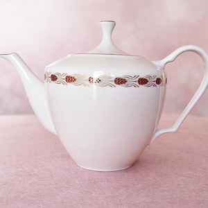 May include: White ceramic teapot with a curved handle and spout. The teapot features a decorative band of brown and gold floral designs around the middle. The lid has a small, rounded finial. The teapot is set against a soft pink background.
