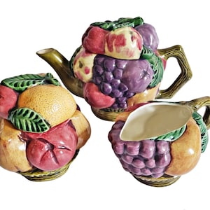 Può includere: Un servizio da tè in ceramica composto da teiera, zuccheriera e lattiera. Il set è decorato con un motivo di frutta colorata, tra cui uva, mele e arance. La teiera ha un manico e un beccuccio a forma di ramo. Lo zuccheriera ha un coperchio.
