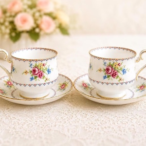 May include: Two white teacups and saucers with floral cross-stitch designs. The cups have gold handles and rims, and the saucers have matching floral patterns. A bouquet of pink roses is in the background.