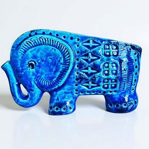 May include: A bright blue ceramic elephant figurine featuring a glossy finish and intricate embossed patterns along its side. The design includes geometric shapes, small circular indentations, and a textured ear detail on a solid blue base.