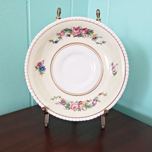 May include: Cream-colored saucer with a floral design and gold trim. The saucer has a white center and a decorative rope-like edge. The floral pattern includes pink, purple, and blue flowers. The saucer is displayed on a brass stand.