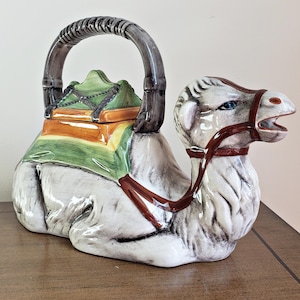 Pode incluir: Uma chaleira de cerâmica em forma de camelo, com uma alça escura e uma tampa. A chaleira é principalmente branca com detalhes em castanho, verde e amarelo. O camelo tem uma rédea castanha e olhos azuis. A chaleira está sobre uma superfície de madeira.