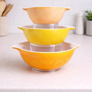 Puede incluir: Una pila de tres cuencos de mezcla Pyrex vintage en tonos naranja y amarillo. Los cuencos tienen bordes y asas blancos. Los cuencos inferior y superior tienen un estampado floral. Los cuencos están sobre una encimera blanca.