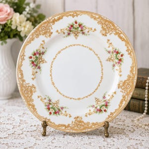 May include: A round white porcelain dinner plate featuring an intricate gold filigree border and a central circular gold pattern. The rim is decorated with four symmetrical floral clusters containing small red, pink, and white flowers.