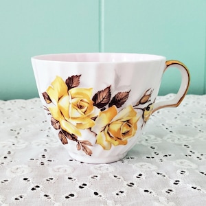 May include: White teacup with a gold handle and rim, adorned with yellow rose floral designs and brown leaves. The teacup is set against a light blue background and rests on a white lace tablecloth.