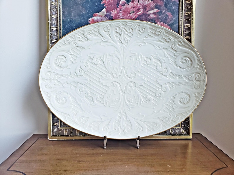 May include: A white ceramic serving platter with a gold rim and an embossed floral design. The platter is on a wooden stand.
