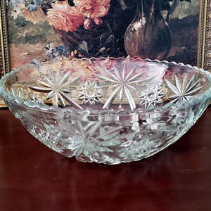 May include: A clear, oval-shaped glass bowl with a starburst pattern. The bowl is placed on a dark wooden surface, with a framed painting of flowers in the background. The bowl's edges have a scalloped design.