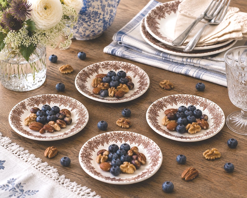 May include: Four small white ceramic plates featuring a brown floral pattern around the rim. Each plate is filled with a mixture of fresh blueberries, walnuts, and almonds. The plates are arranged on a wooden surface alongside scattered nuts.