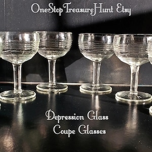Peut inclure: Six verres à coupe en verre transparent avec un motif côtelé. Les verres sont disposés en deux rangées de trois. Le texte "Depression Glass Coupe Glasses" est imprimé sous les verres.